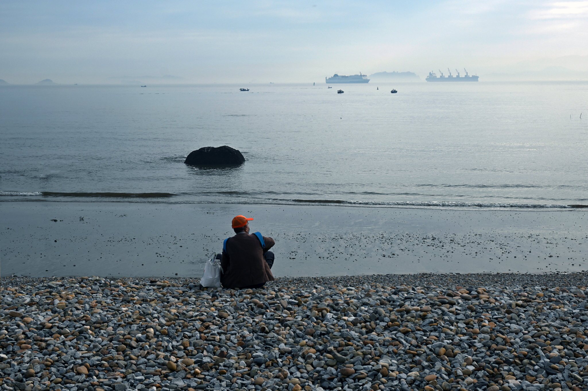 一人坐在卵石滩上向远处眺望平静的海面